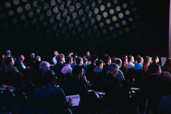 Audience seated in a dimly lit venue, focused on a presentation or speaker.